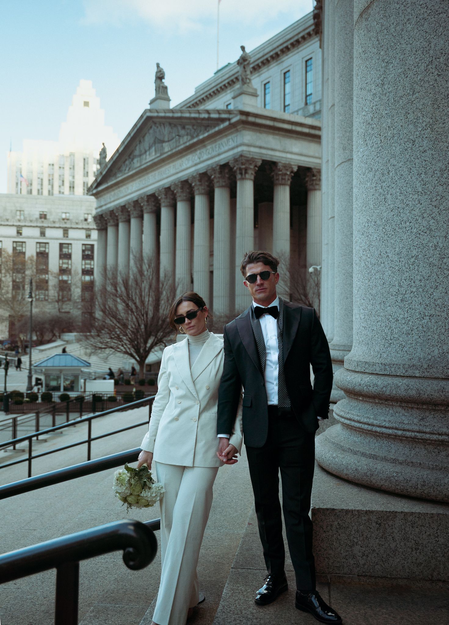 A couple in formal wear poses between classic stone columns of a grand building.