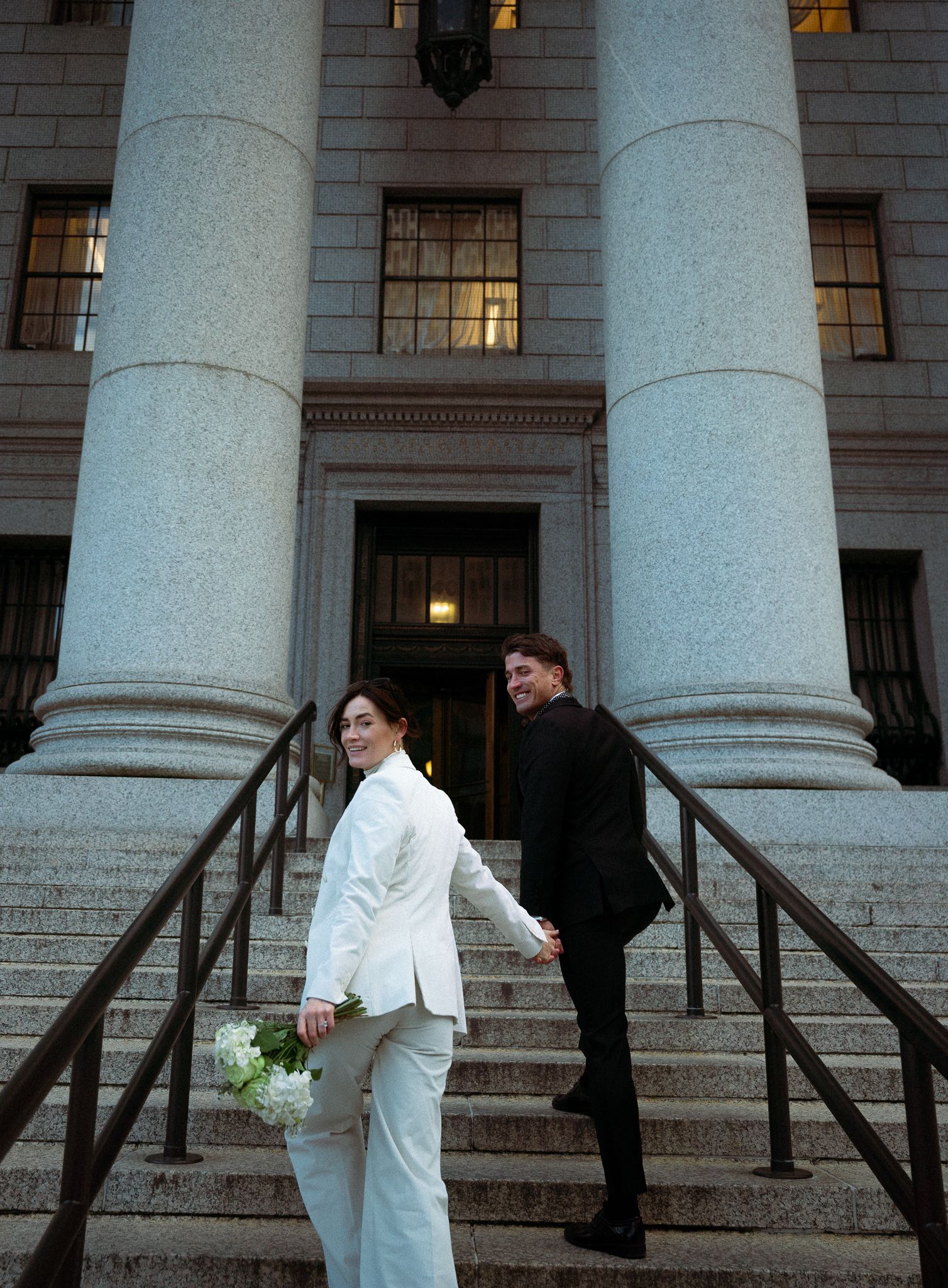 A couple in contrasting black and white attire poses on the steps of a columned building.