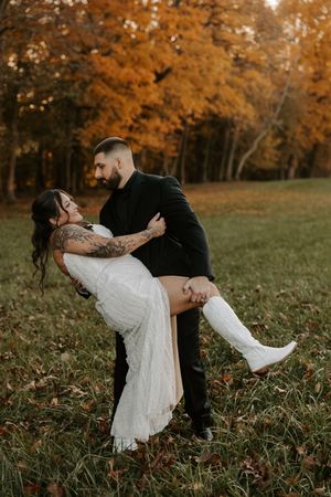 Playful wedding couple dips in a grassy meadow surrounded by fall trees with golden leaves.