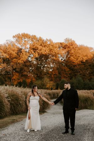 Wedding couple walks along a gravel path bordered by tall ornamental grasses and autumn trees.