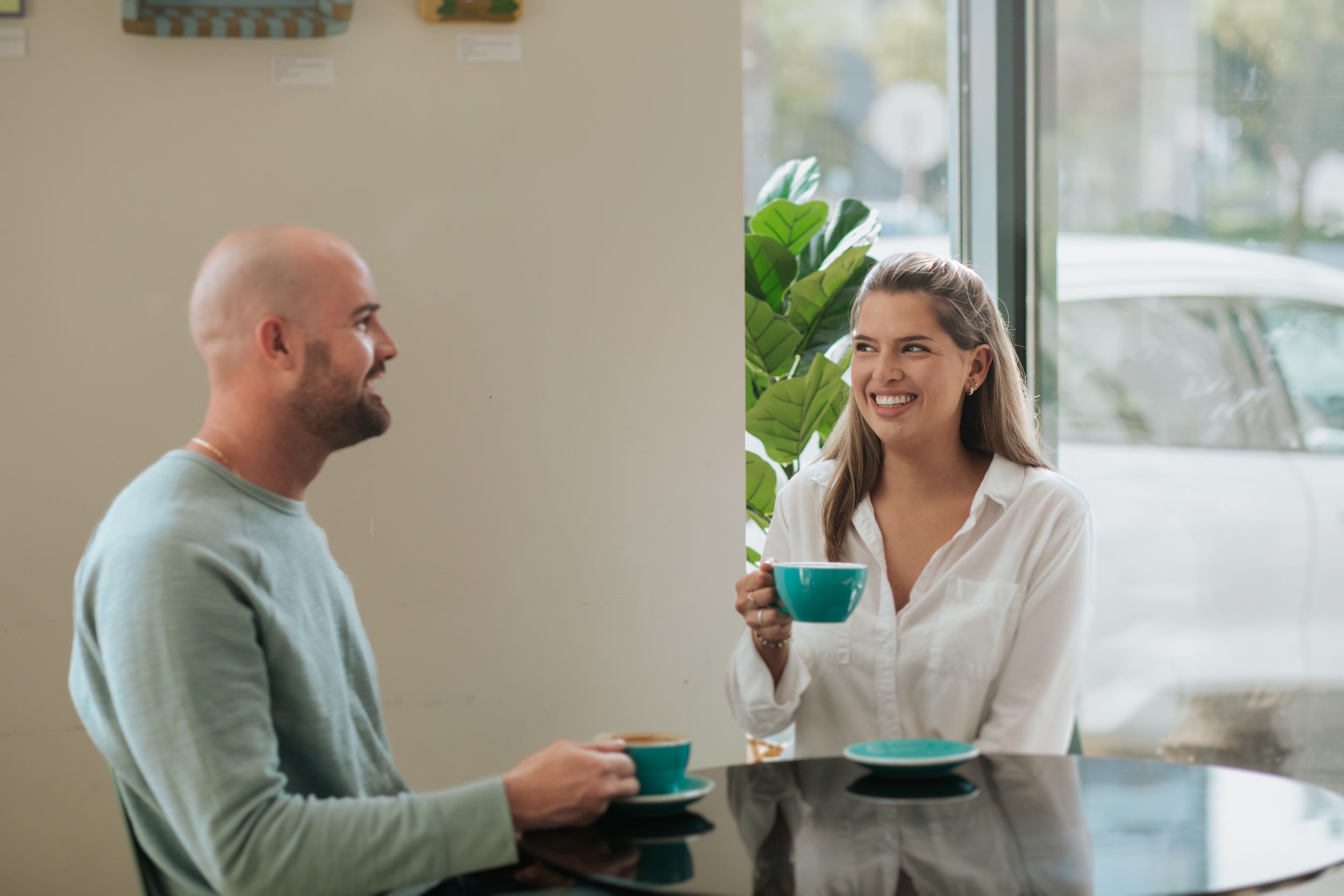 Two people enjoying coffee and conversation while sitting at a table near a window in a bright room.