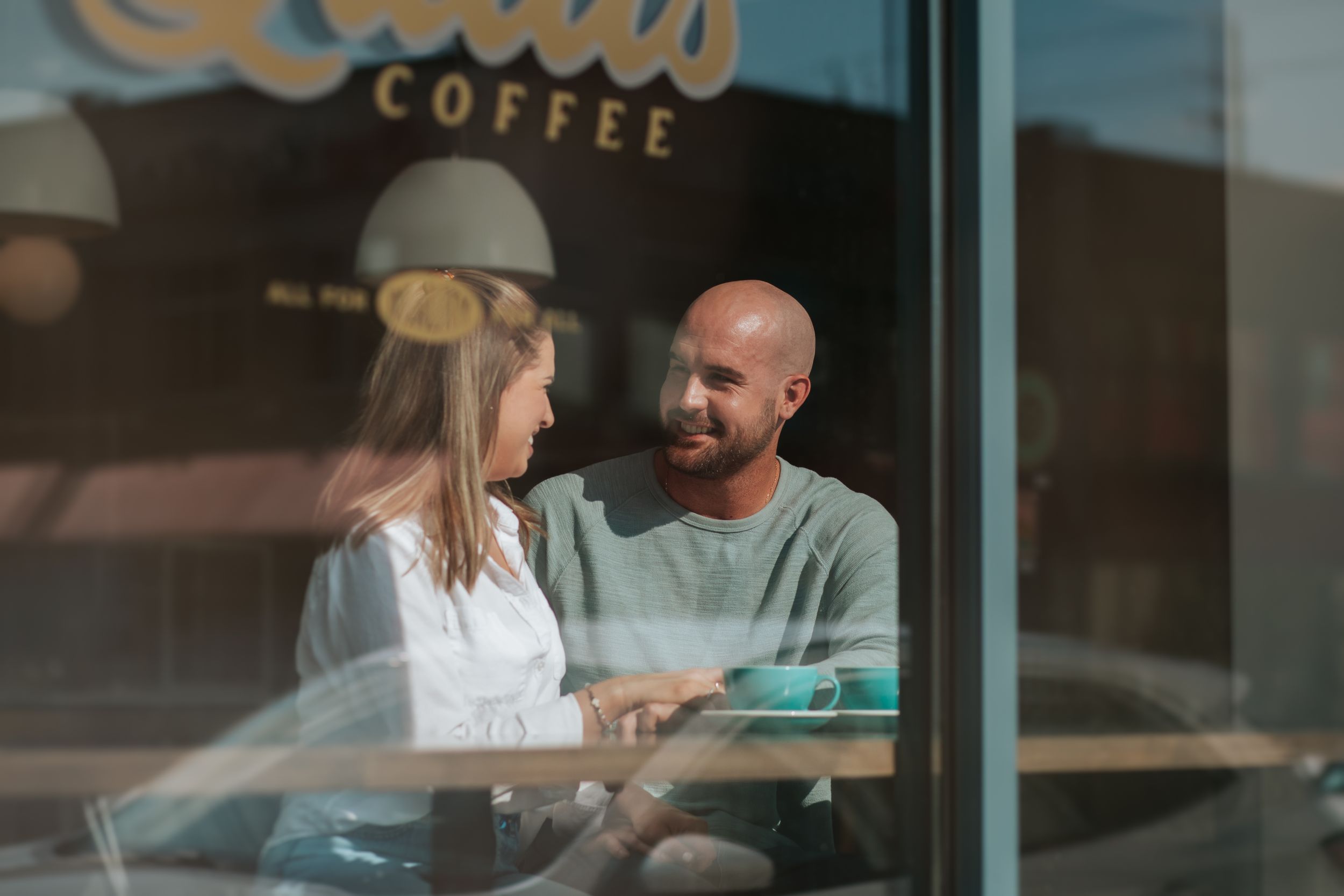 Two people share a conversation at a coffee shop table seen through a window with reflections.