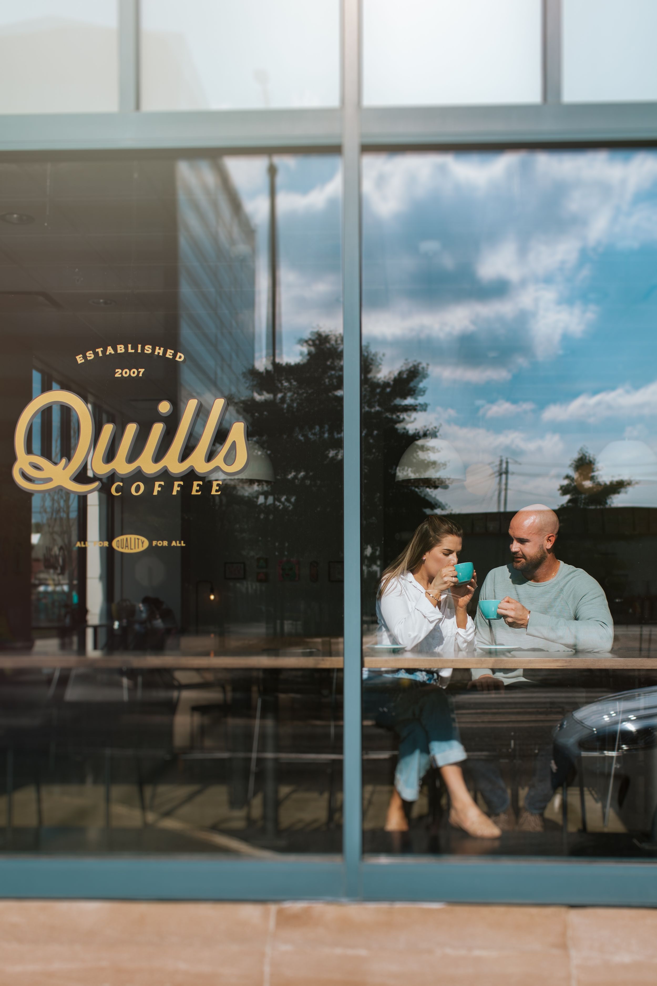 Through a coffee shop window, a couple sits at a table enjoying conversation and coffee together in warm afternoon light.