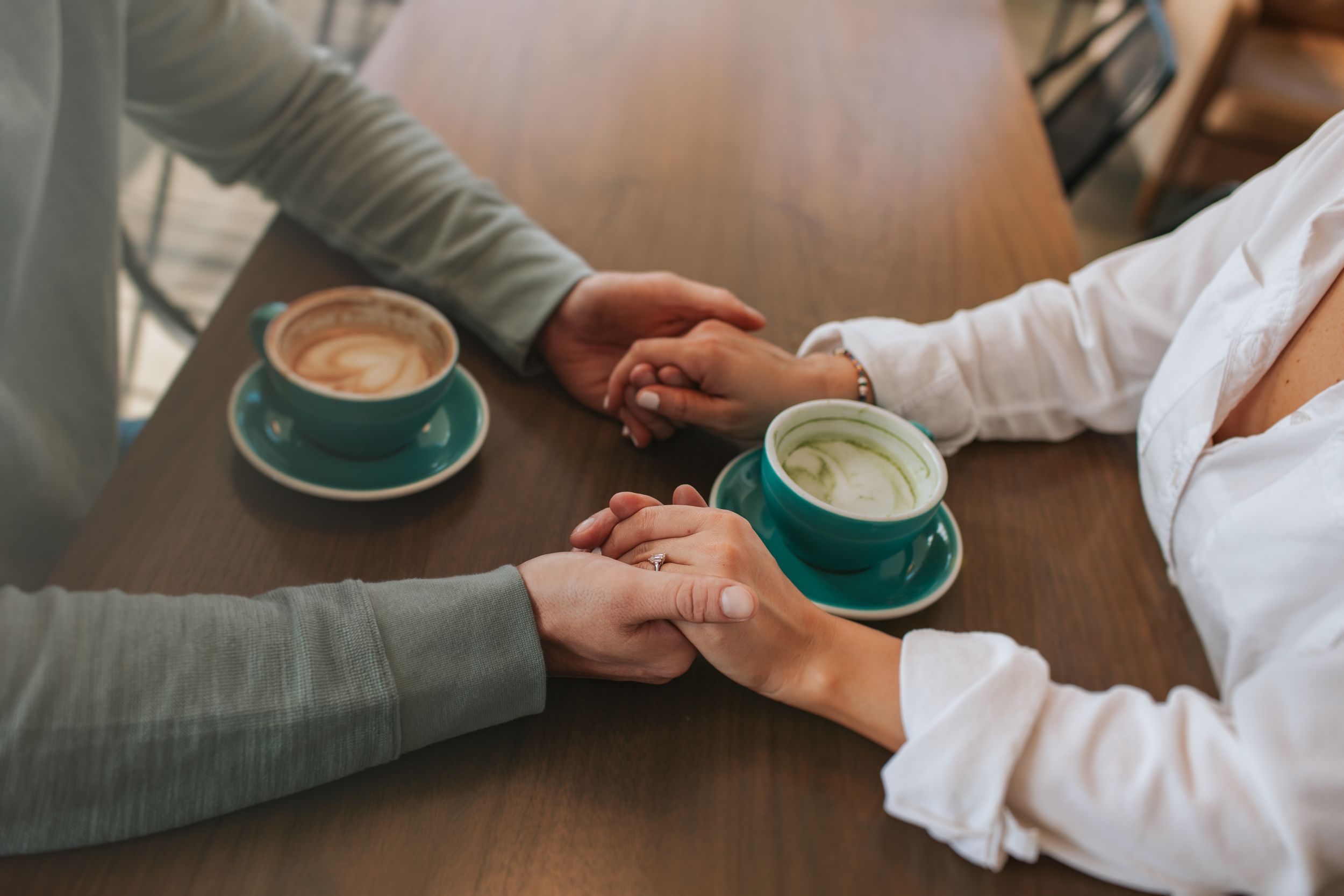 Two people hold hands across a wooden table with turquoise and white coffee cups during an intimate cafe moment.
