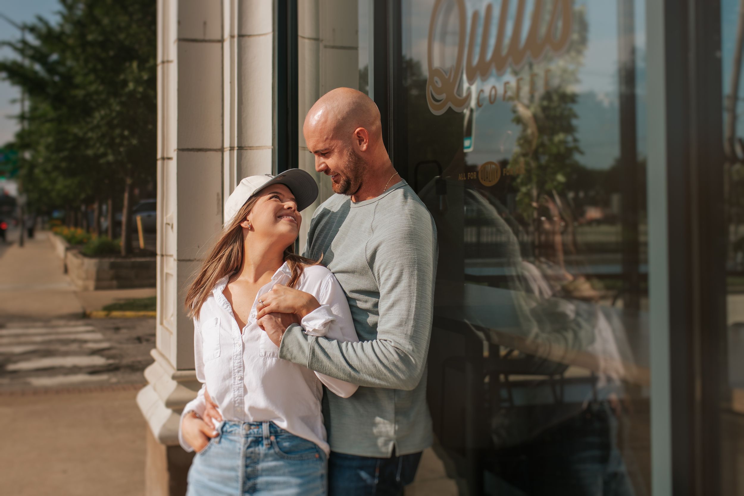 A couple shares an intimate moment while standing together outside a storefront window on a sunny afternoon.