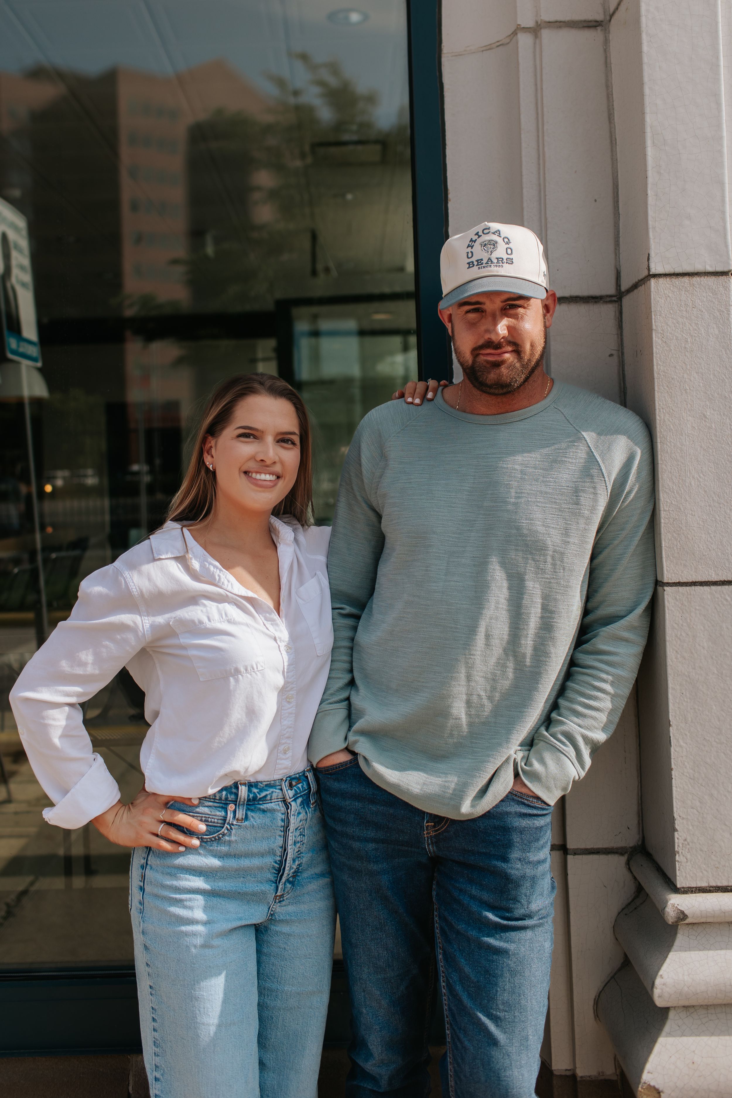 Two people stand casually against a white building exterior wearing jeans, a white shirt and gray sweater.