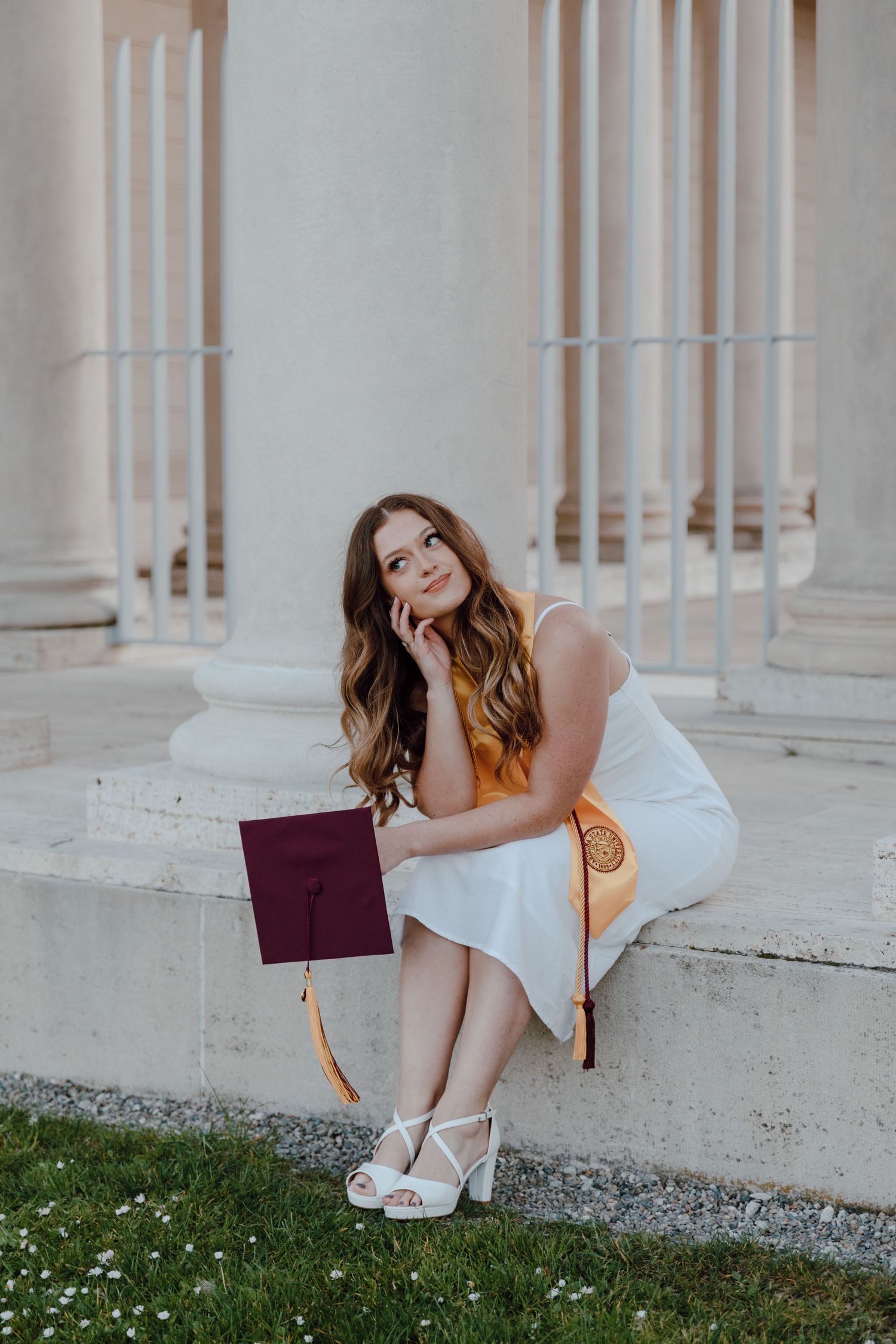 Graduate in white dress poses thoughtfully with diploma against classical columns and grass. Taken by Bay Area Graduation Photographer.