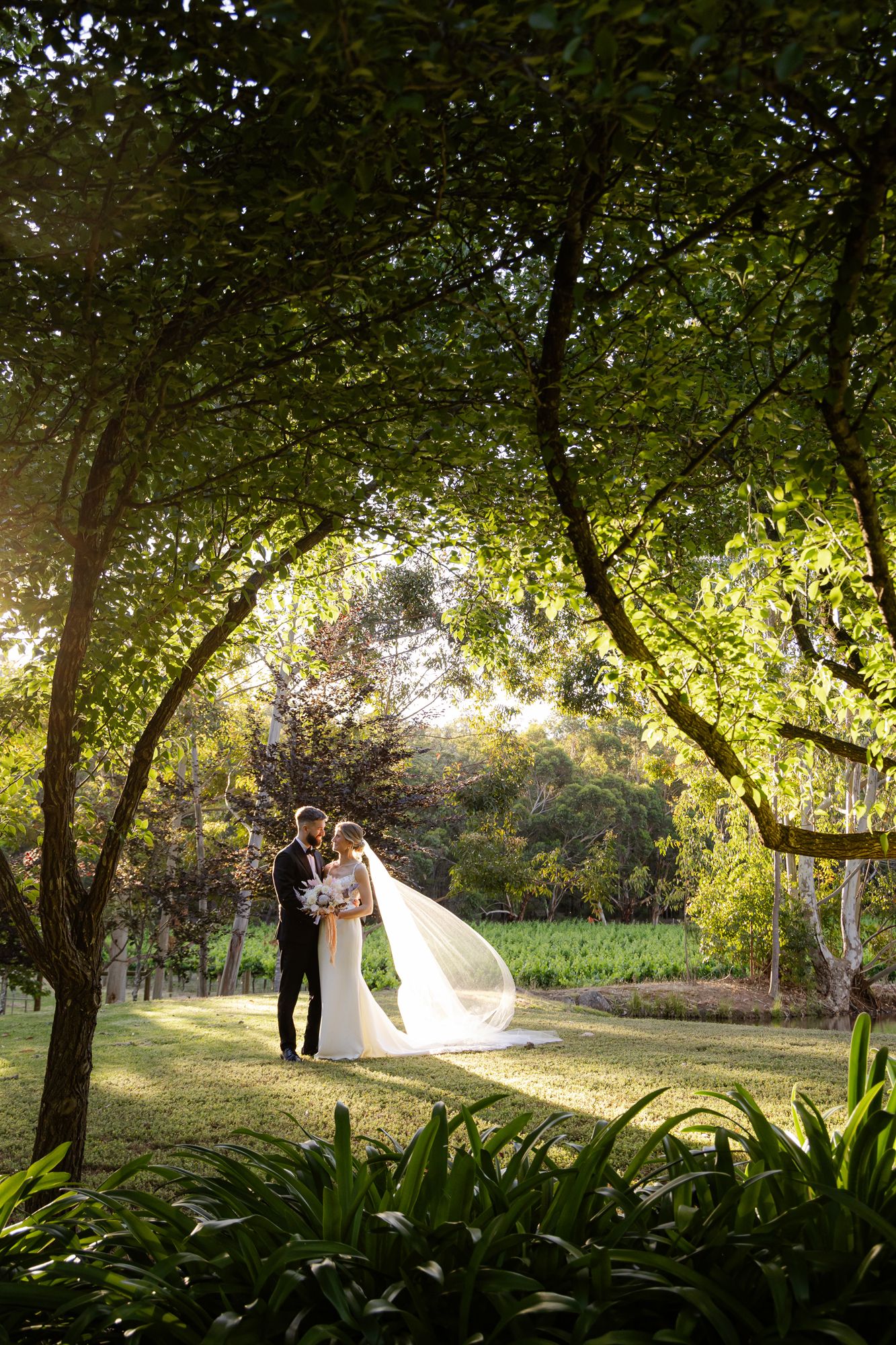 A bride and groom stand on stone steps against a bright blue sky at sunset, with architectural details in view.