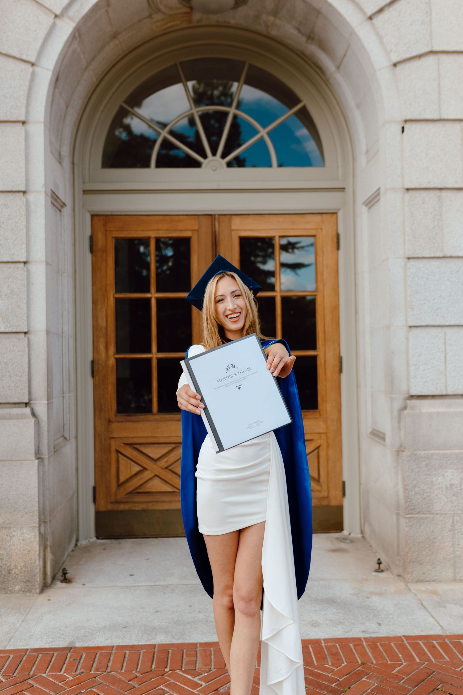 UC Berkeley graduate poses outside on the campus for her grad photoshoot. She wears a cap and gown and holds her master's thesis.