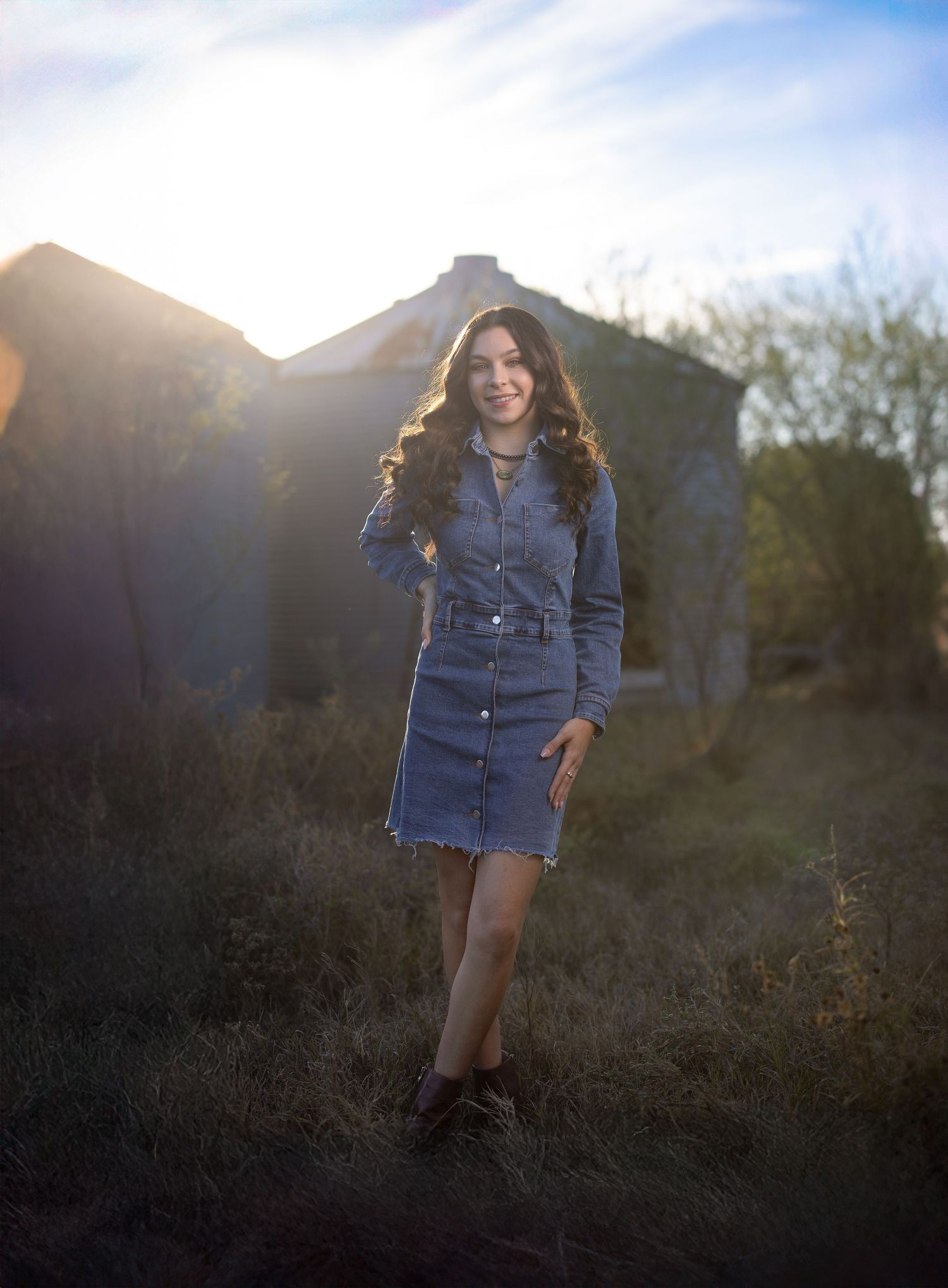 A young woman in a denim dress poses outdoors during golden hour with sunlight creating a dreamy backlit effect.