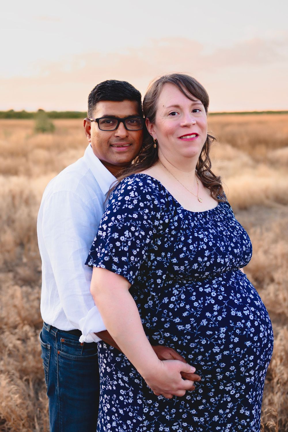 A couple in love poses together in a field at sunset, with the woman wearing a blue floral dress.