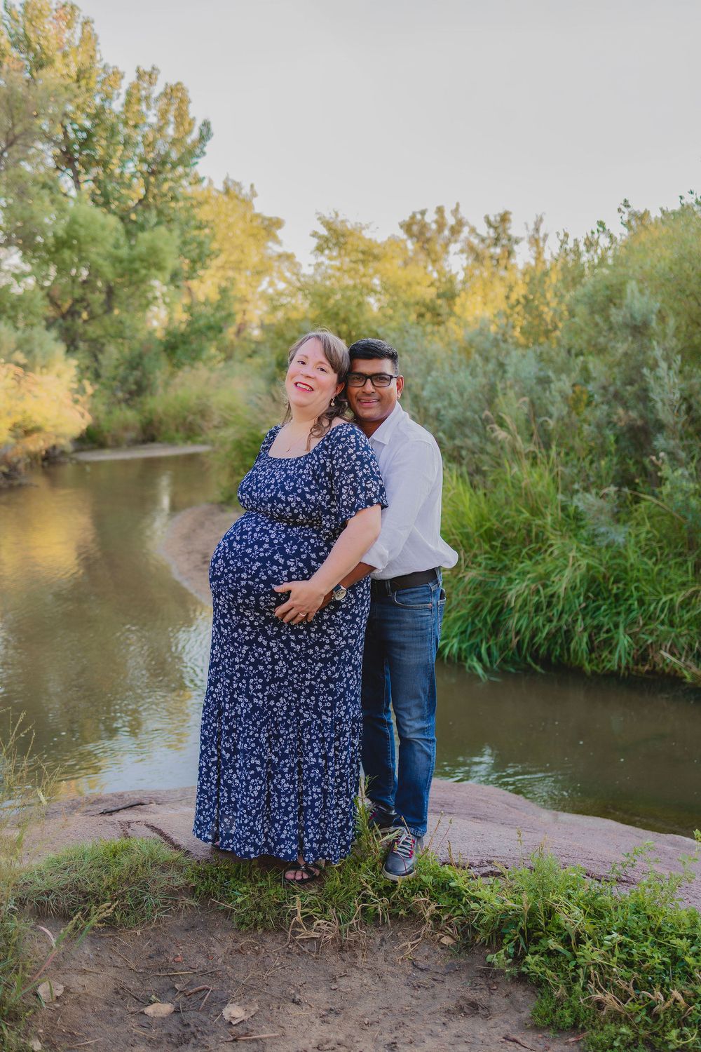 A couple shares an intimate moment by a river surrounded by autumn foliage and greenery.  Maternity photoshoot.