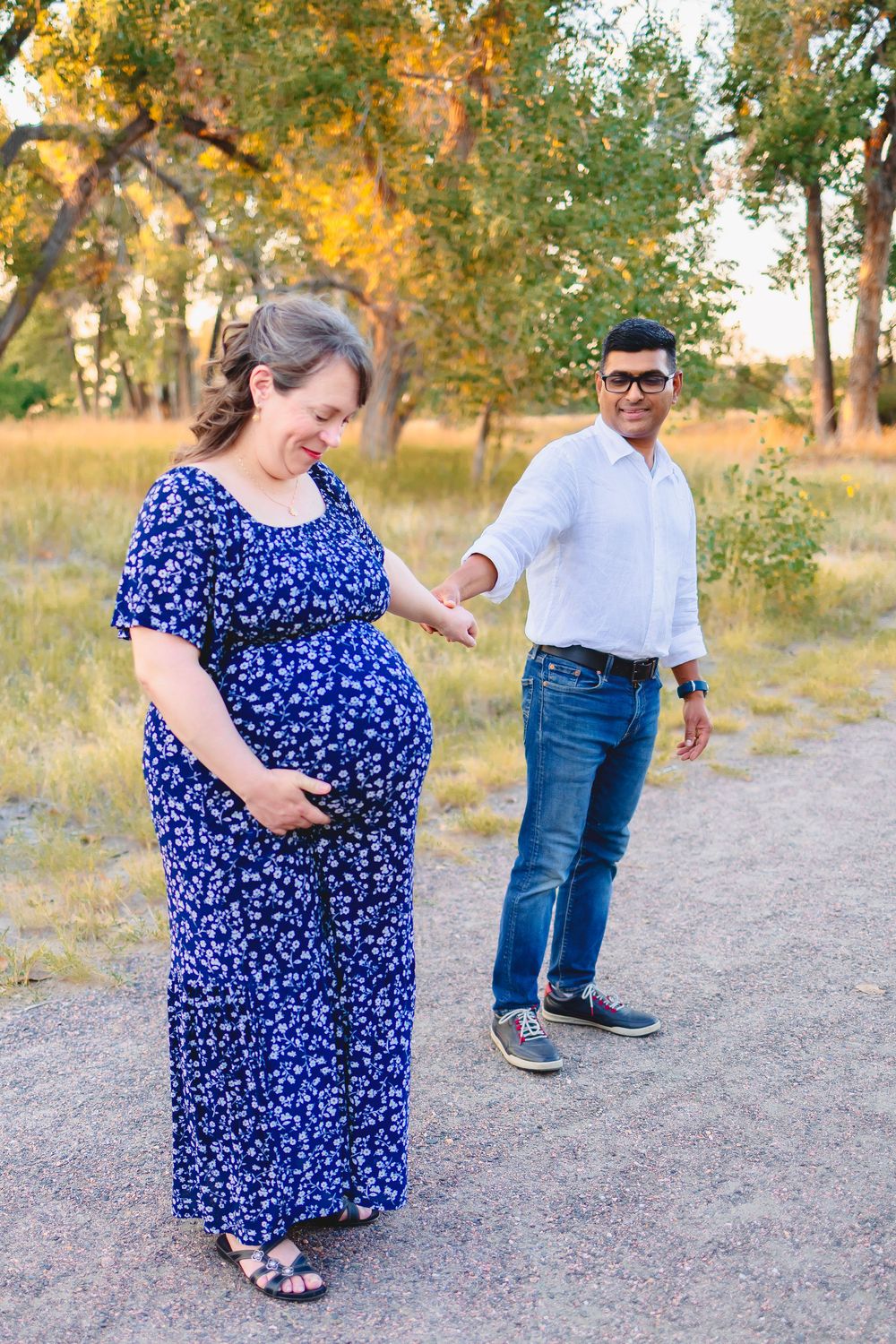 A couple shares a tender moment on a path surrounded by autumn trees during an outdoor maternity photoshoot.  Woman is looking down at bump