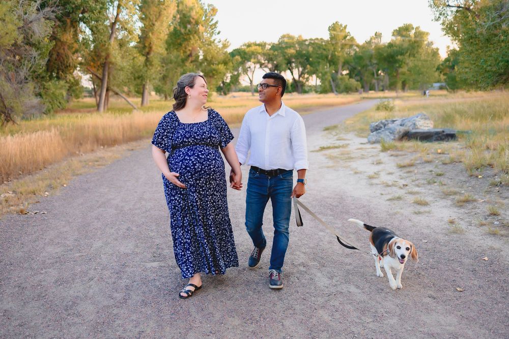 A couple walks their small dog along a dirt path surrounded by green trees on a sunny day.  Maternity photoshoot