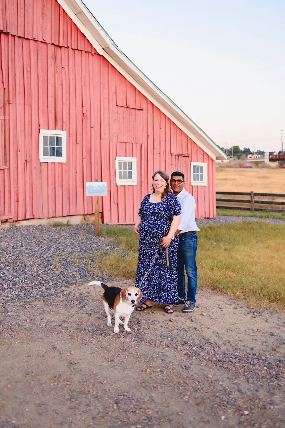 A couple stands near a rustic red barn in a rural setting with a dog on a gravel path at sunset. Maternity photoshoot