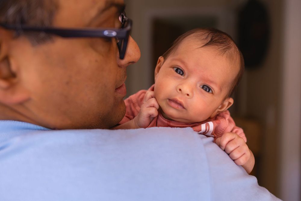 A small infant peeks over an adult's shoulder while being held in a blue shirt.