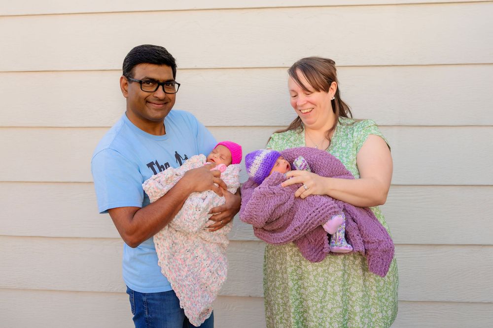 Two people stand outside holding babies wrapped in pink and purple blankets against a beige wall.