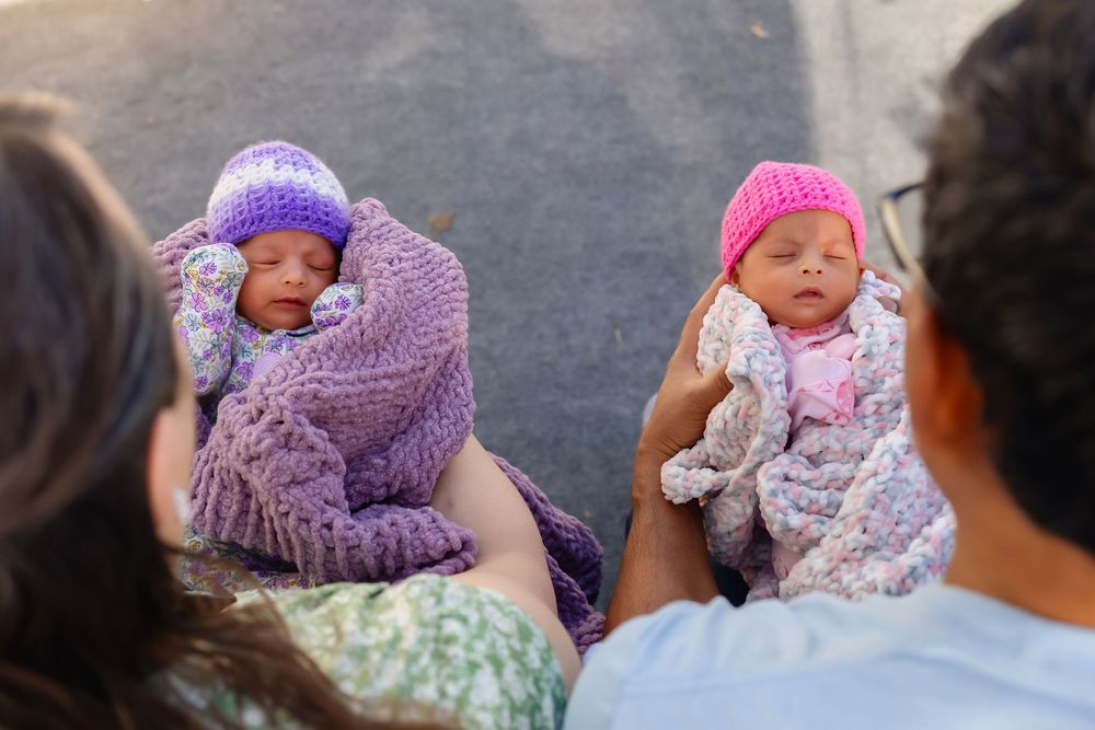 Two sleeping newborn twins wrapped in purple and pink knit blankets and hats held by parents outdoors.