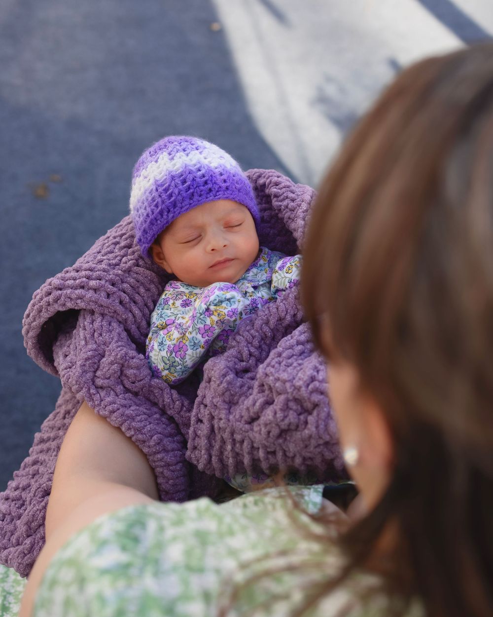 A sleeping baby wrapped in a purple knit blanket wears a purple and white hat with floral accents.