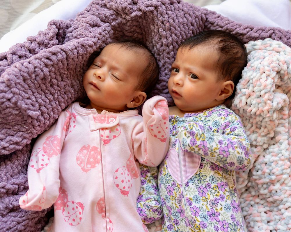 Two babies wearing floral sleepwear lie close together on soft bedding with a purple knit blanket.
