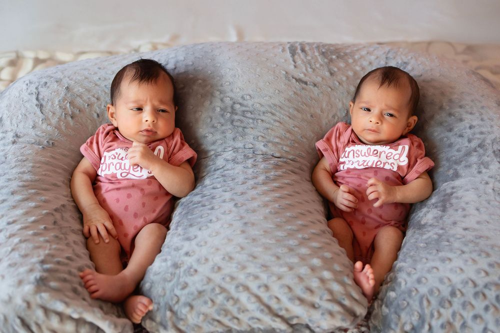 Two babies wearing matching pink outfits lie side by side on a plush gray blanket.