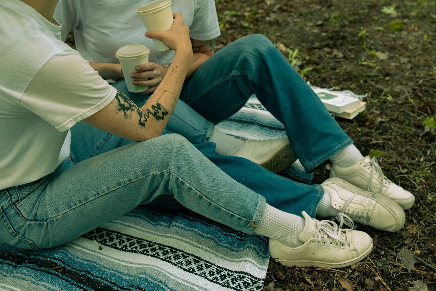 Two people in casual denim and sneakers sitting on a patterned blanket outdoors enjoying a relaxed moment.