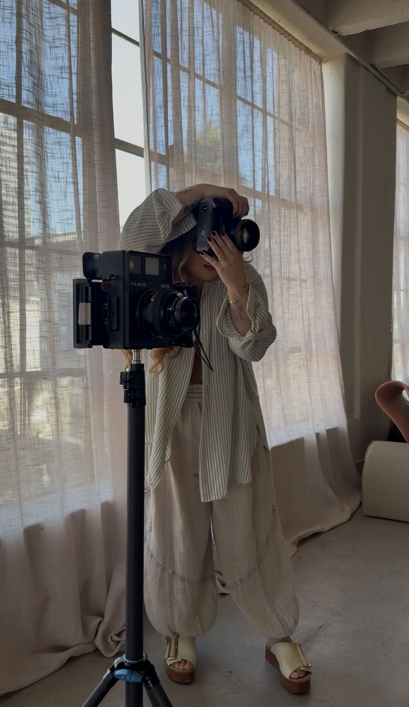 A photographer in light beige linen clothing works with a professional camera mounted on a tripod near sheer curtained windows.