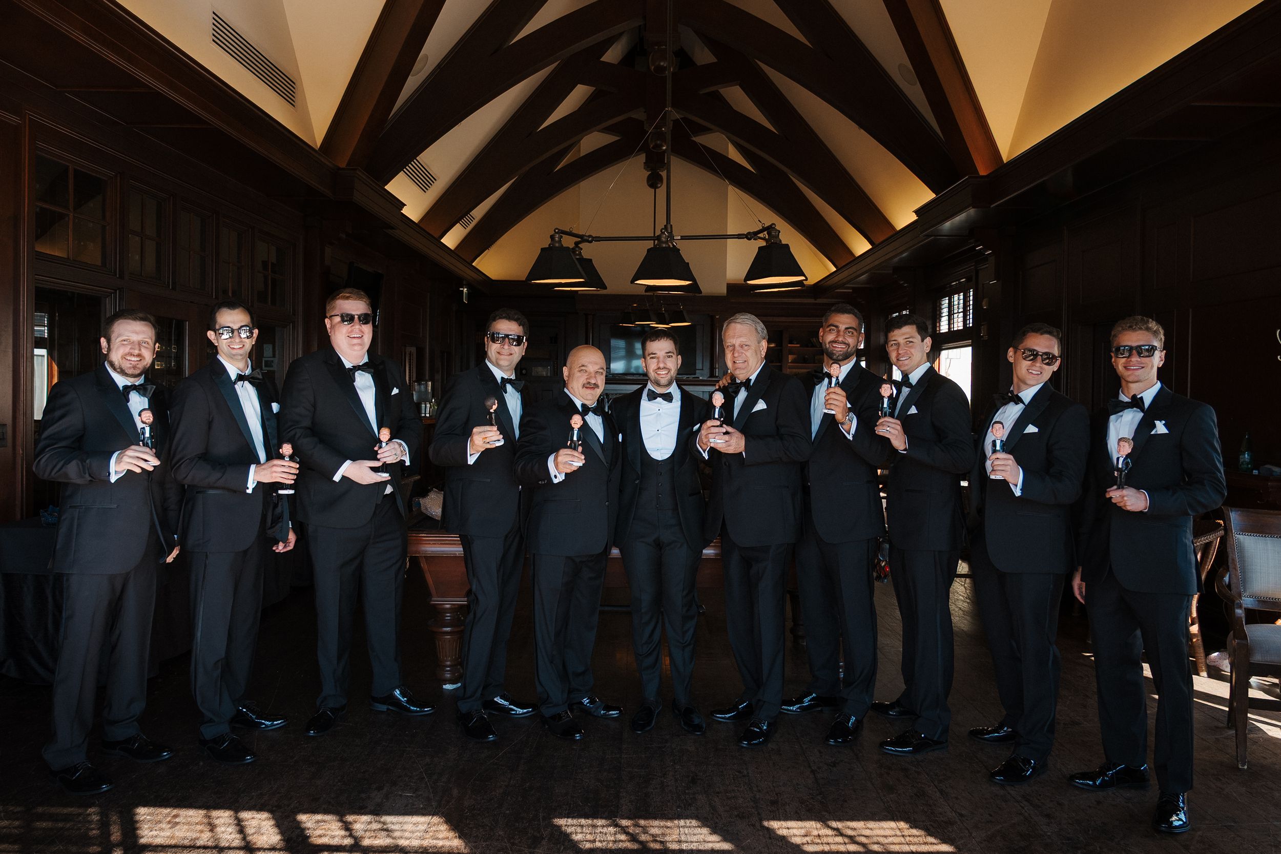 Groomsmen in black tuxedos pose in line against a dark wooden backdrop.