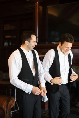 Two men in formal black vests and white shirts share a laugh while looking at a mobile phone.