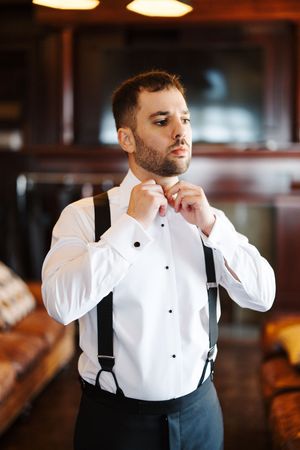 A person in formal attire adjusts their bowtie while wearing a crisp white dress shirt and black suspenders.