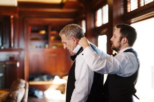 A groomsman helps adjust a bow tie on someone in a formal white dress shirt in an elegant wood-paneled room.