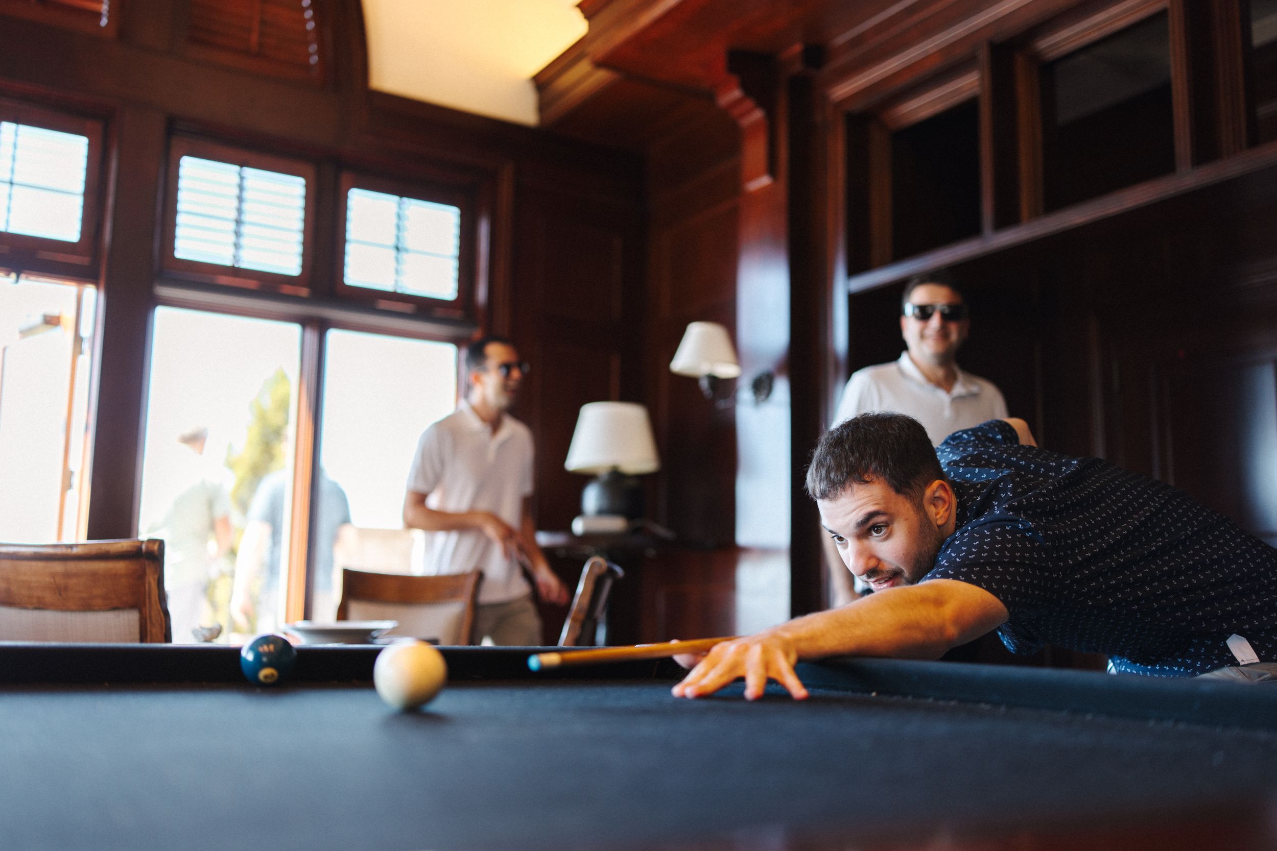 A person plays a game of pool in a dimly lit billiards room with dark wood paneling and warm lighting.