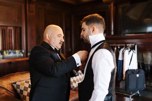 Two men in formal attire interact in an elegant room with wooden paneling and hanging garments in the background.