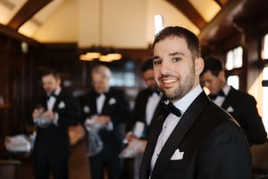 Groomsmen in black tuxedos gather at an indoor venue with warm lighting and elegant decor.