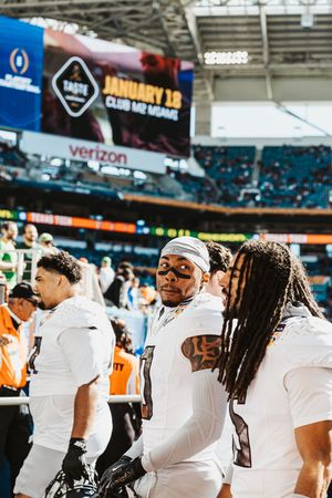 Football team members in white uniforms stand together on the sidelines of a stadium field.
