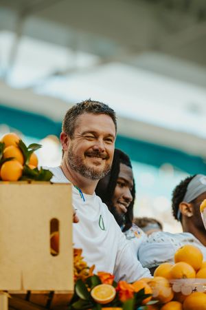 A smiling person in a white uniform poses with fresh oranges and citrus fruits in a bright setting.