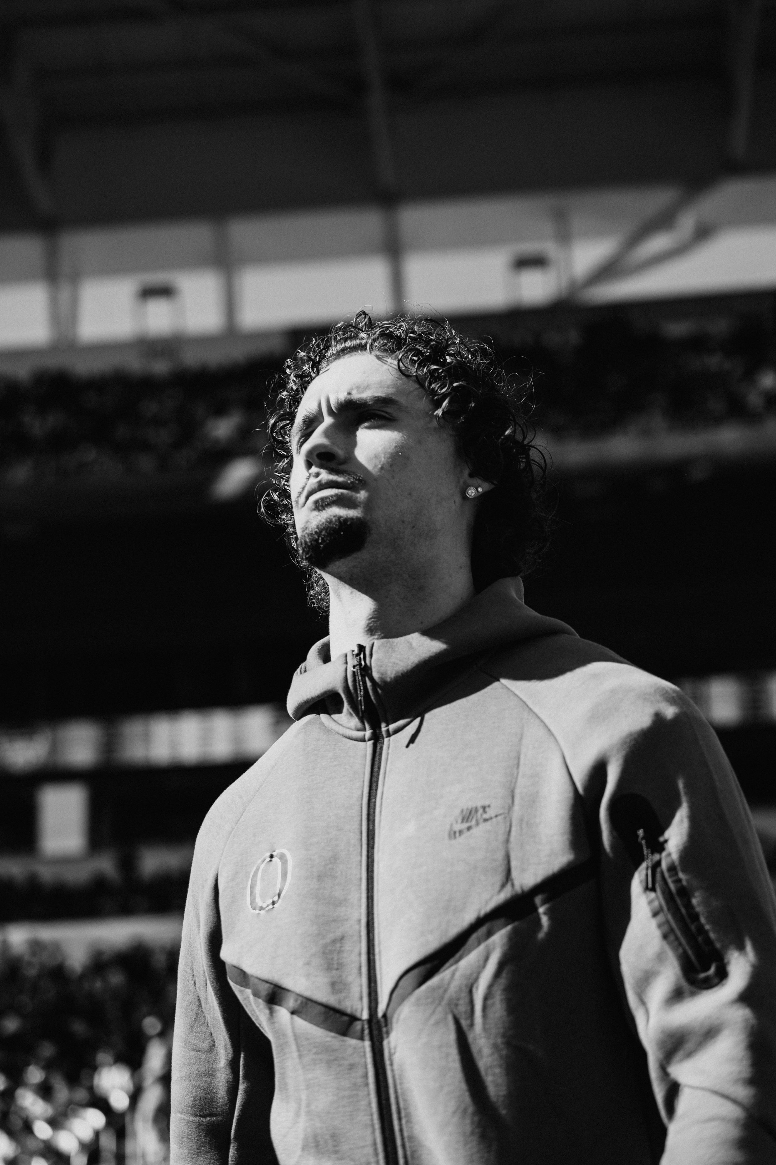 Black and white portrait of person in athletic jacket looking upward at stadium venue.