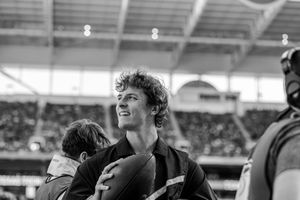 Black and white photo of a smiling player holding a rugby ball in a stadium with spectators in the background.