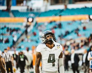 Players in white uniforms stand on a football field with teal stadium seats visible.