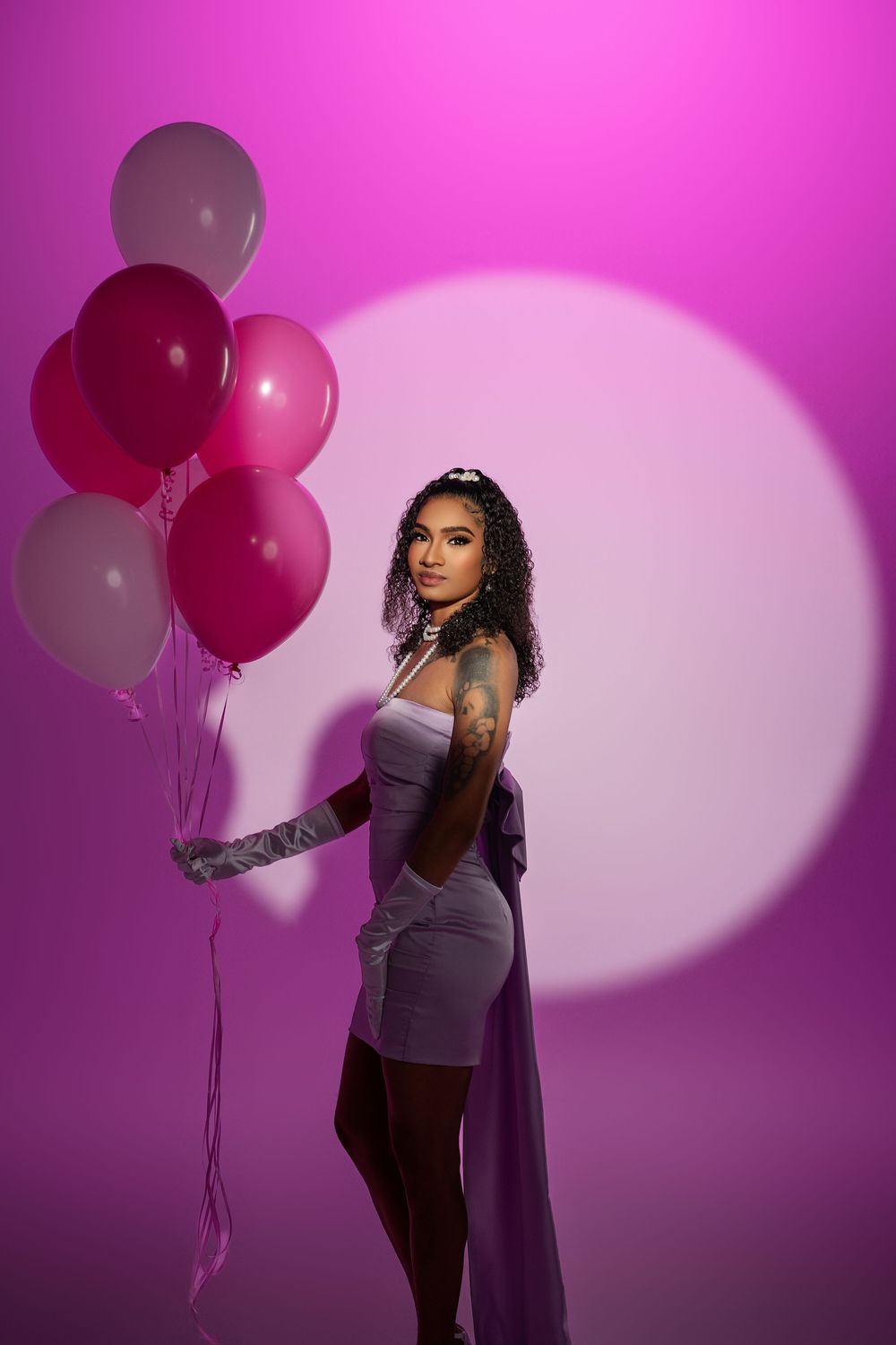Fashion photoshoot against pink backdrop with balloons featuring purple outfit and dramatic lighting.