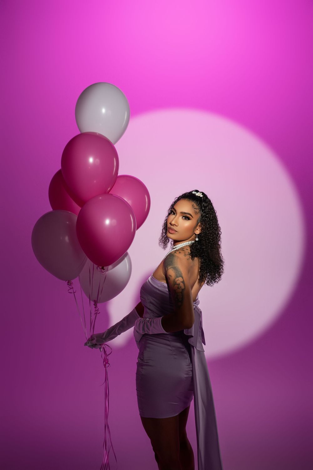 Fashion model in purple dress poses with pink and white balloons against vibrant magenta backdrop with spotlight effect.