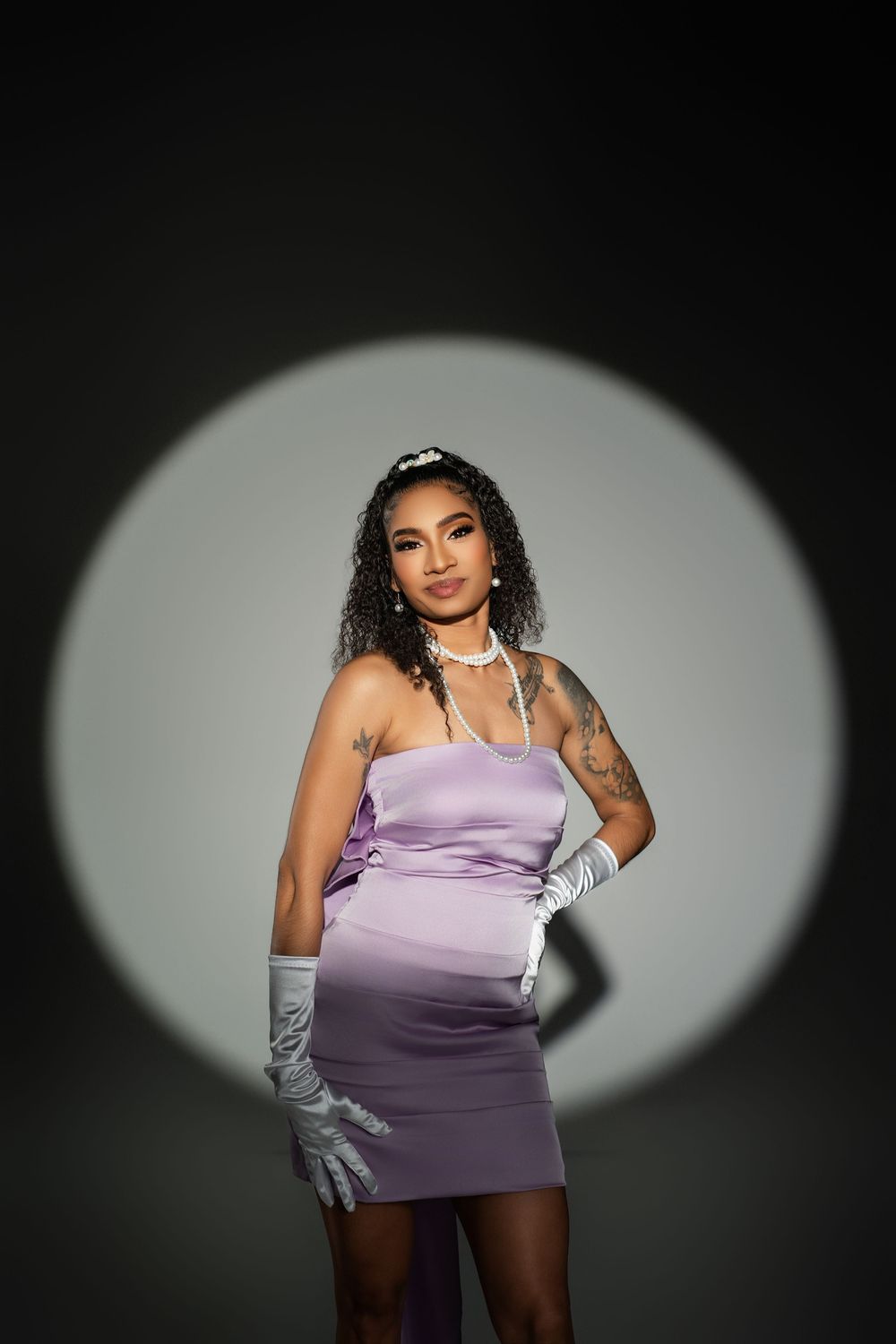 Fashion model in purple dress and silver gloves poses against dark backdrop with dramatic spotlight.