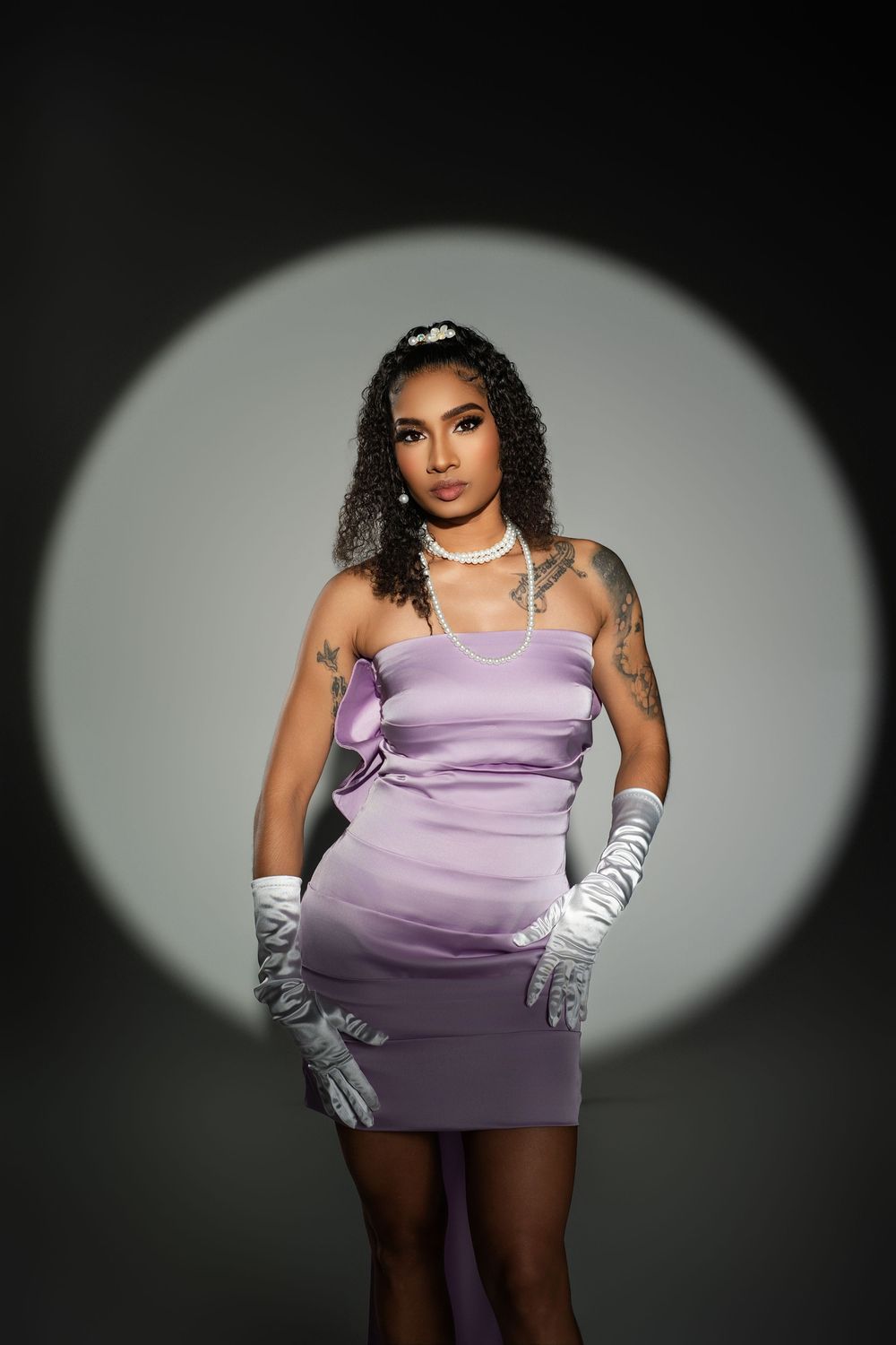 Fashion model poses in lavender dress and silver gloves against dramatic spotlight backdrop.