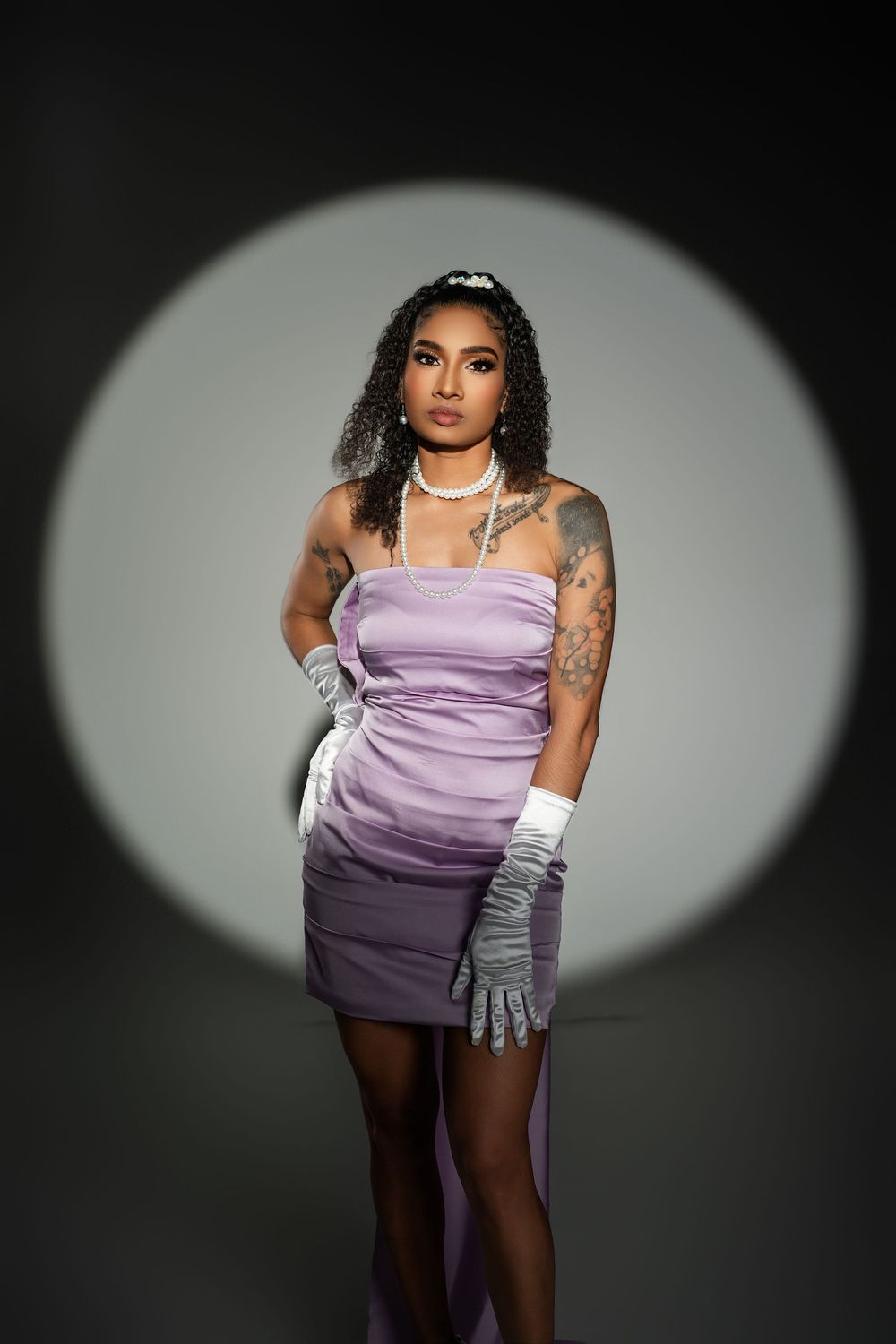 Fashion model in purple dress and silver gloves poses dramatically against dark backdrop with spotlight effect.