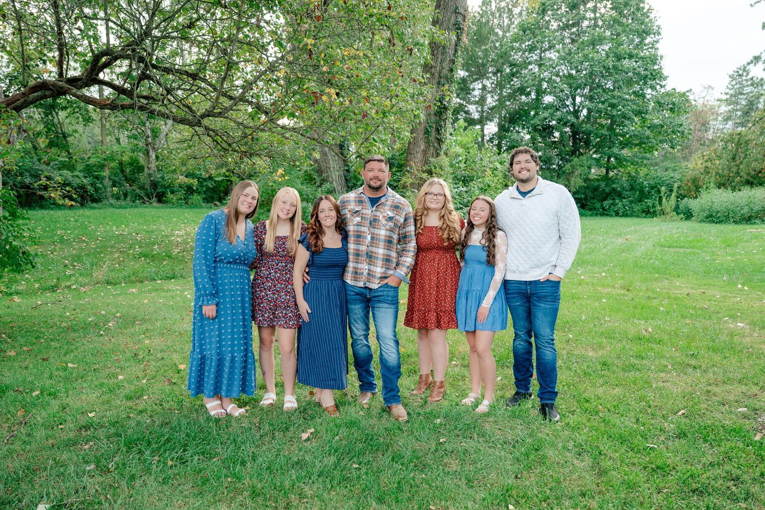 A group stands together on a grassy lawn wearing coordinated casual outfits in blue and earth tones.