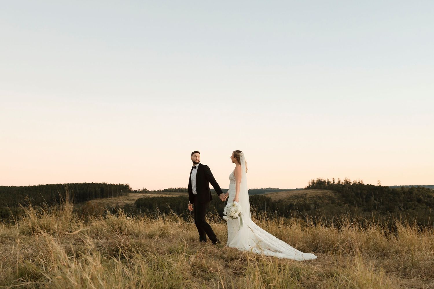 A couple in wedding attire stands together on a hilltop during golden hour with tall grass and trees in the distance.