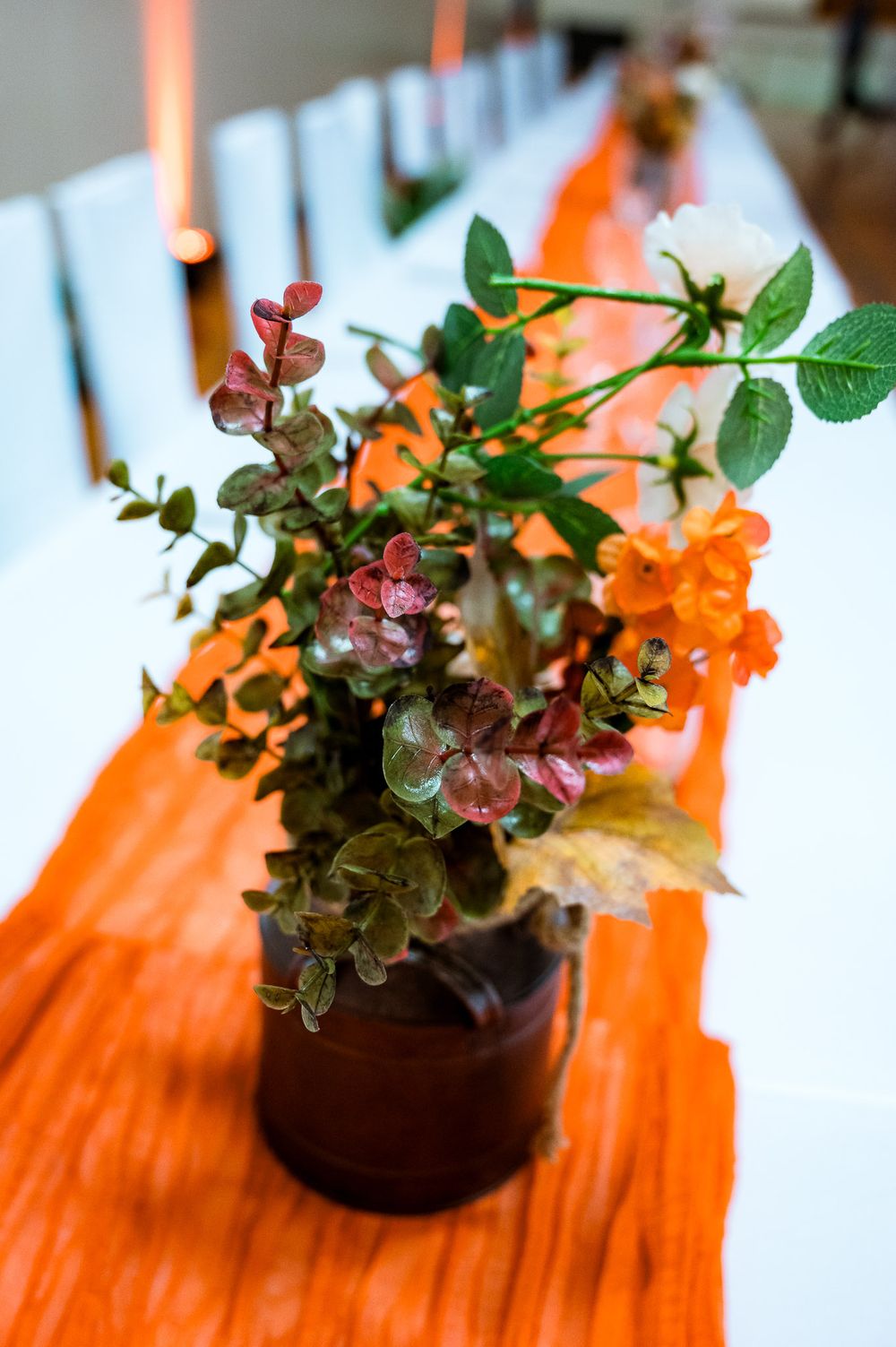 A potted plant with burgundy and green foliage sits on an orange table runner with white candles in the background.