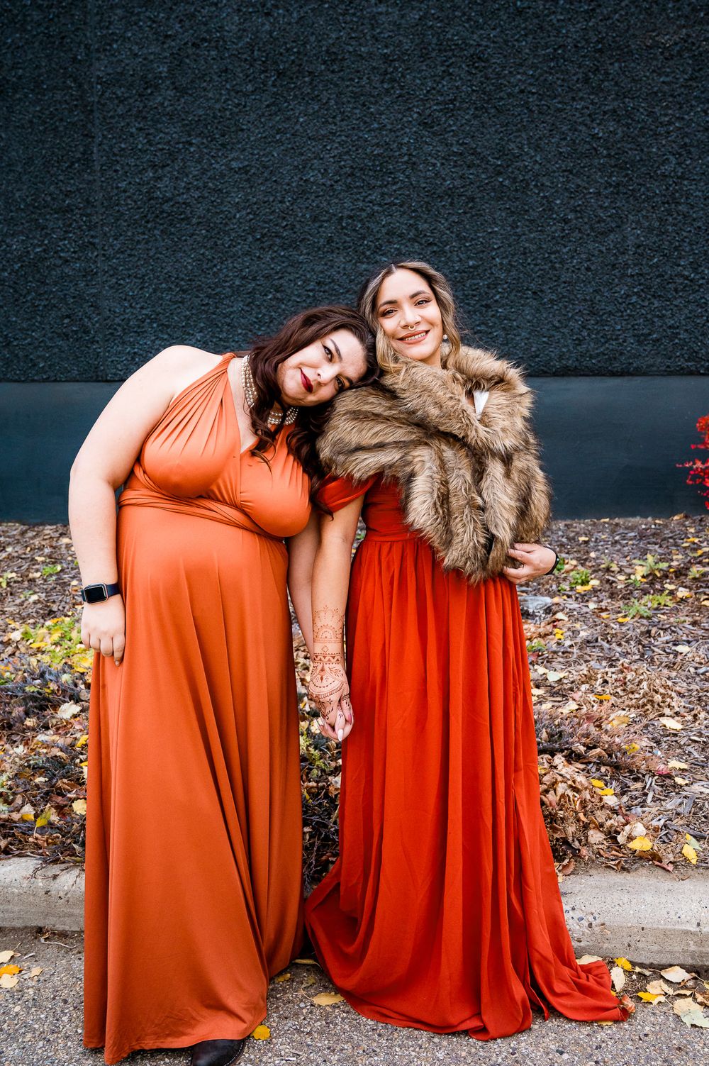 Two people in flowing orange and rust-colored dresses pose playfully against a dark wall with autumn leaves on the ground.