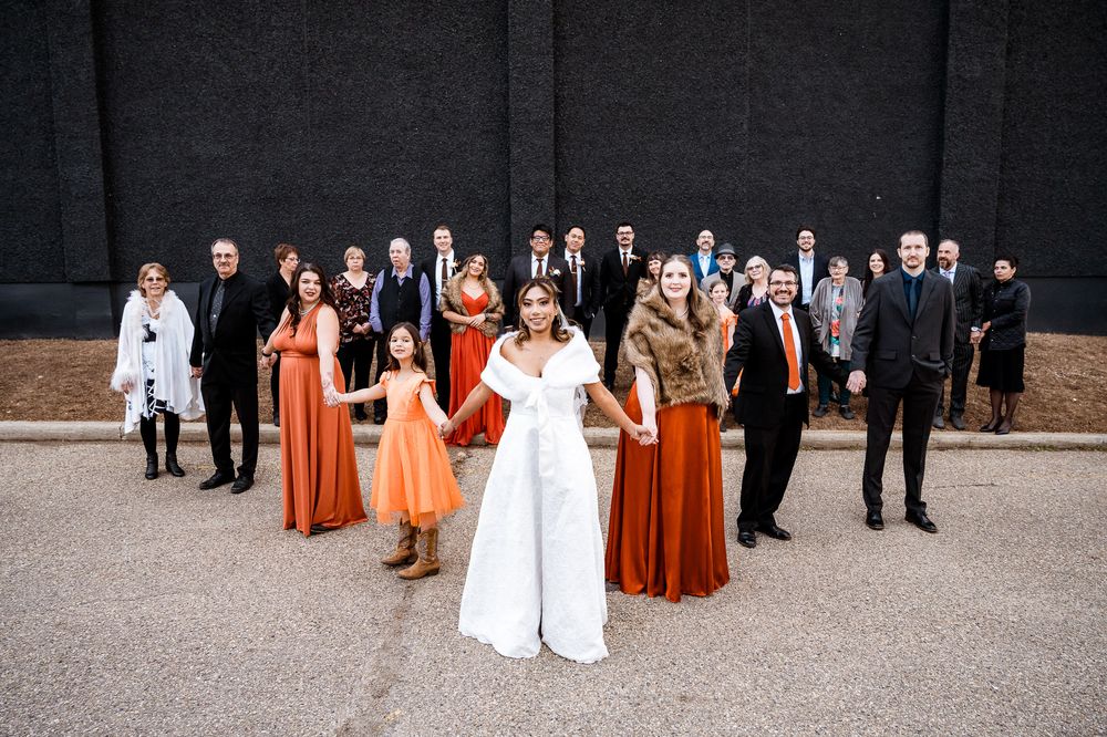 Wedding party members in formal attire gather for a group photo against a dark wall outdoors.