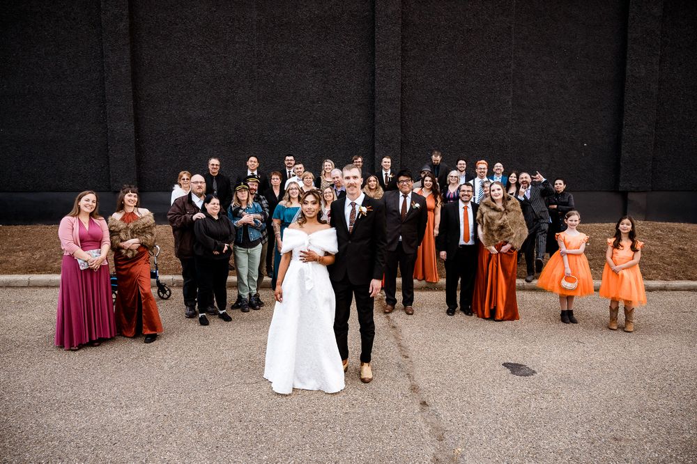 Wedding party group photo with bridesmaids in orange dresses and groomsmen in black suits gathered outdoors.
